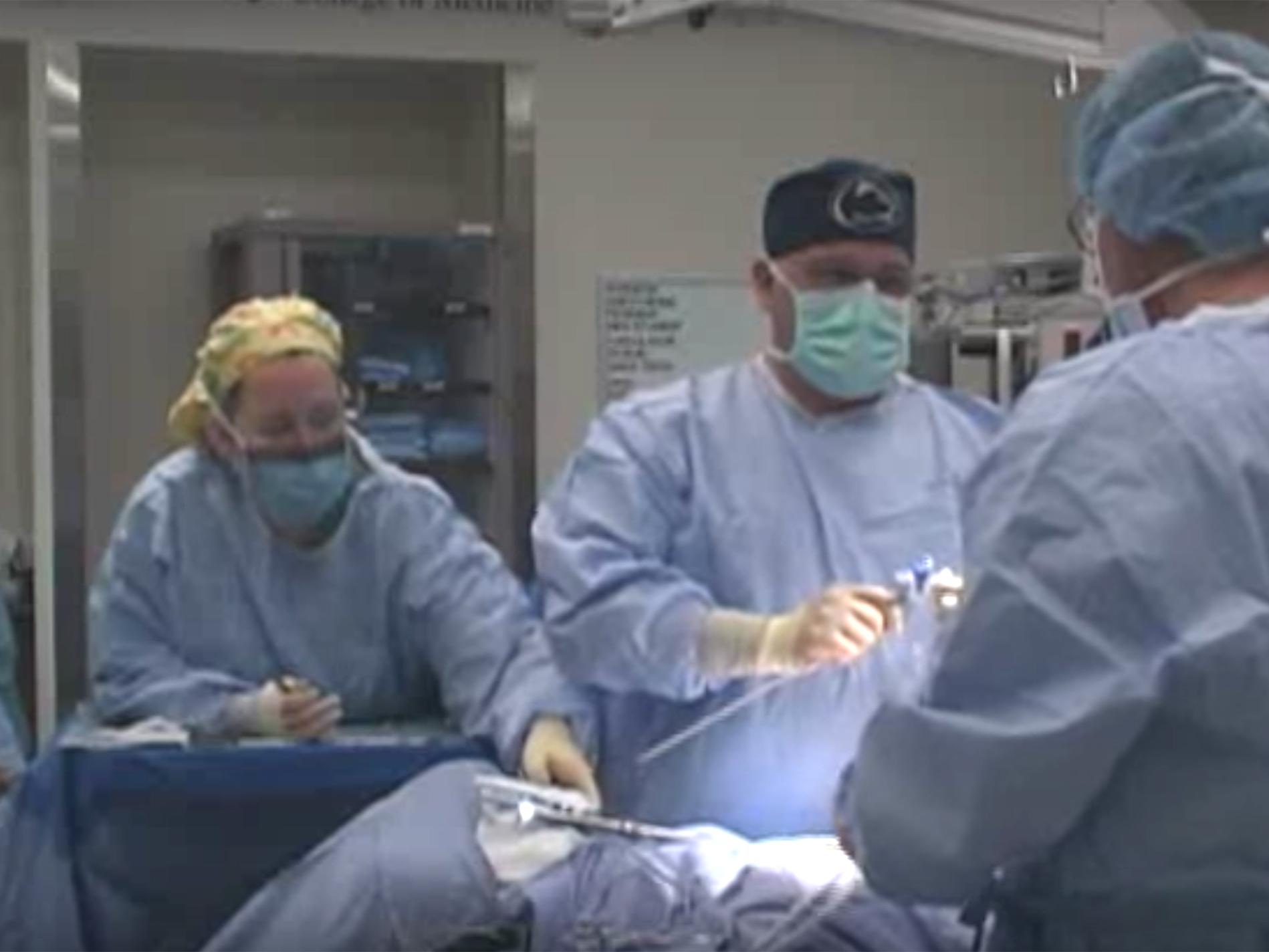 Dr. Gerald Harkins holds an operating tool and looks at an assistant during a procedure featured on Surgery Live! He wears a surgical gown, gloves, cap and mask. A nurse in a gown and mask next to him readies surgical instruments on a tray.