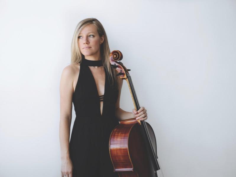 A woman stands and holds her cello in her left hand while she looks to her right.