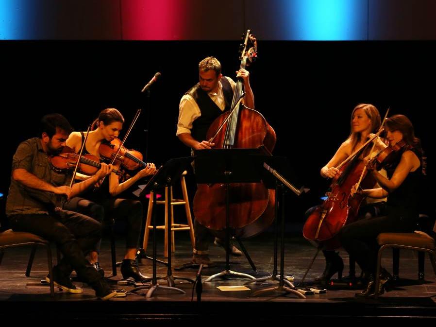 The musicians of Sybarite5, four of them seated while the double bassist stands, perform on stage.