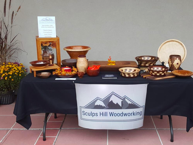 Photo of craft fair vendor table with black table cloth, sign that reads "Sculps Hill Woodworking," and several examples of the bowls and charcuterie boards they offer.