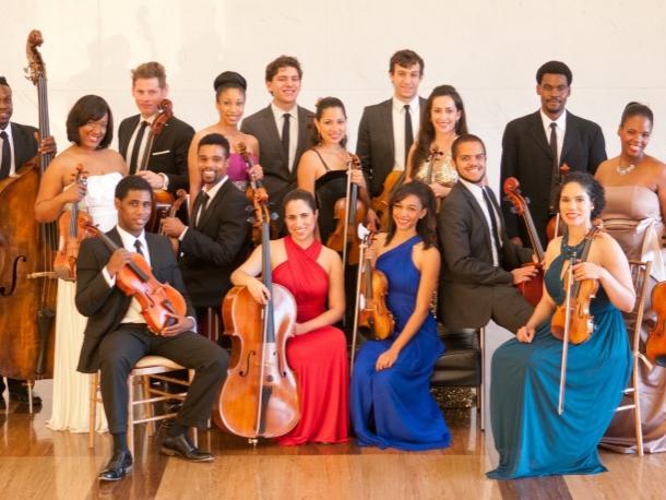 A diverse group of formally dressed musicians hold their instruments and smile for the photographer.