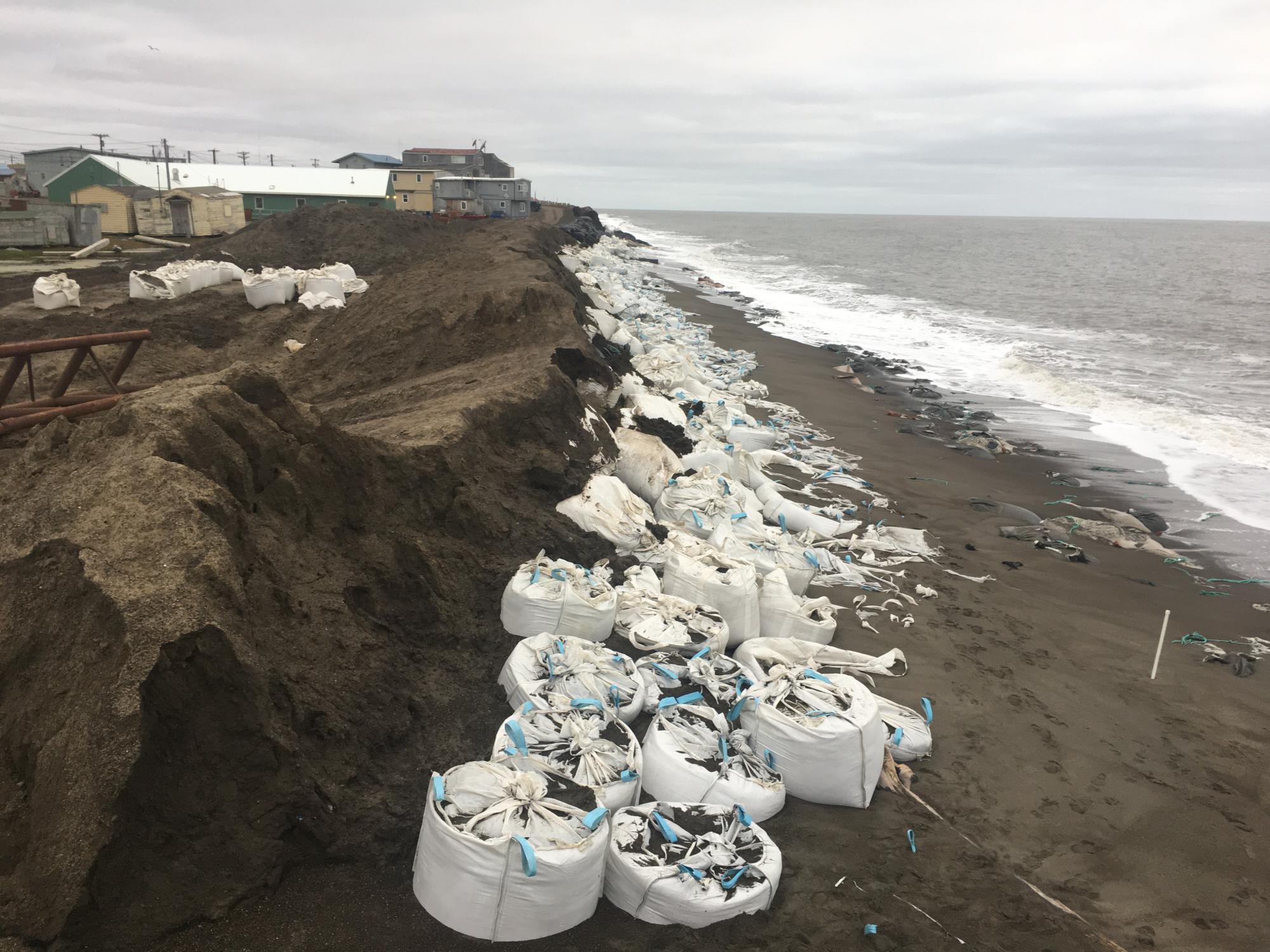 White bags line an eroded coastline, with buildings in the background. Ripped bags trail across the beach into the ocean. 