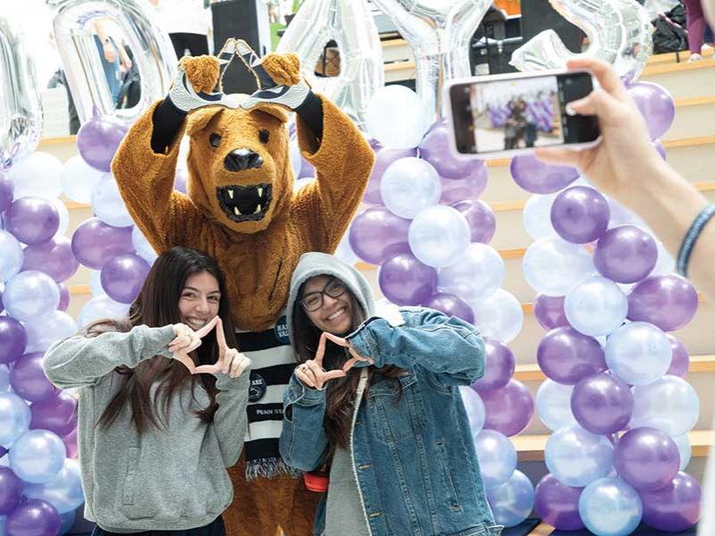 The Nittany Lion poses for a photo with two students in front of a balloon background.