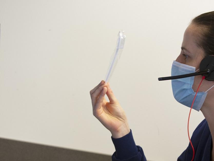 A person in profile wears a surgical mask and holds a toothbrush up to a computer monitor. The person holding the toothbrush also wears a headset.