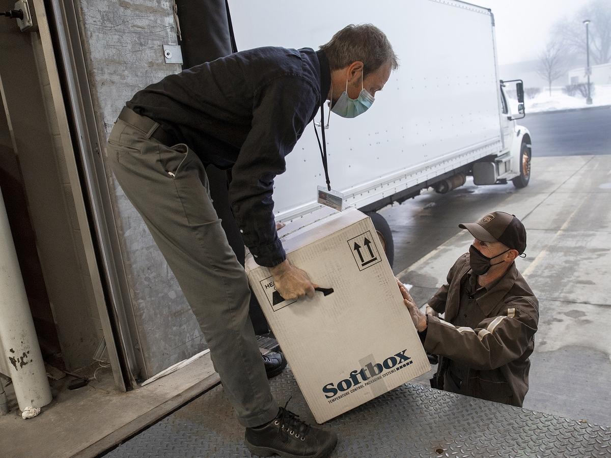 A man on a loading dock wears a mask and stoops to pick up a cardboard box placed on the dock by a man in a ballcap in a surgical mask.