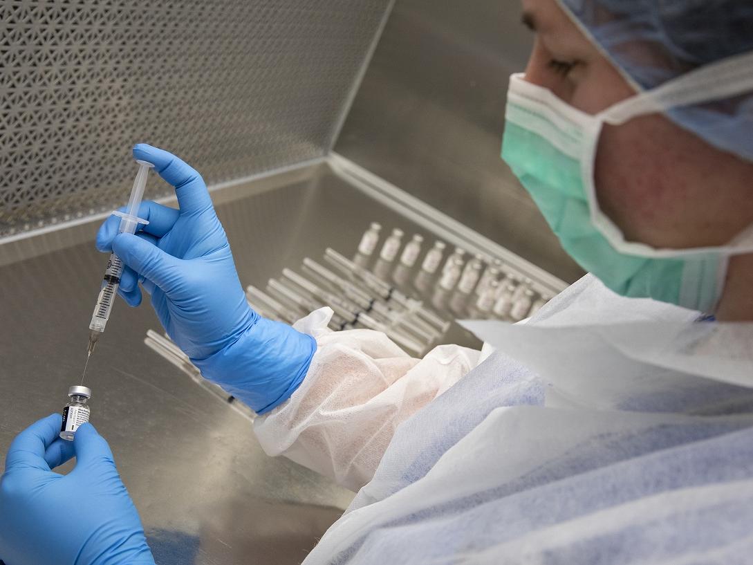 A woman in personal protective equipment inserts a syringe into a vial.