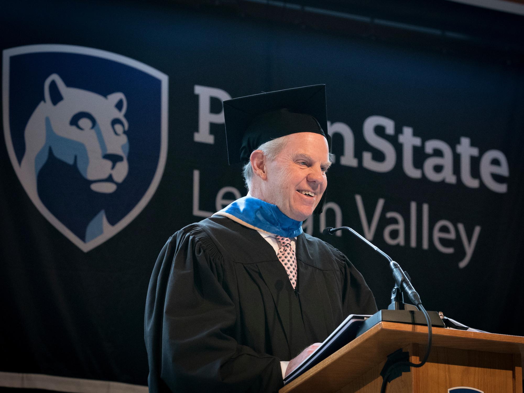 man in cap and gown delivering remarks at podium