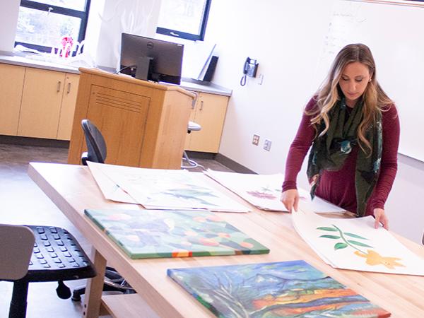 Lecturer in Art, Corianne Thompson stands at large art table in an art studio and looks at student artwork