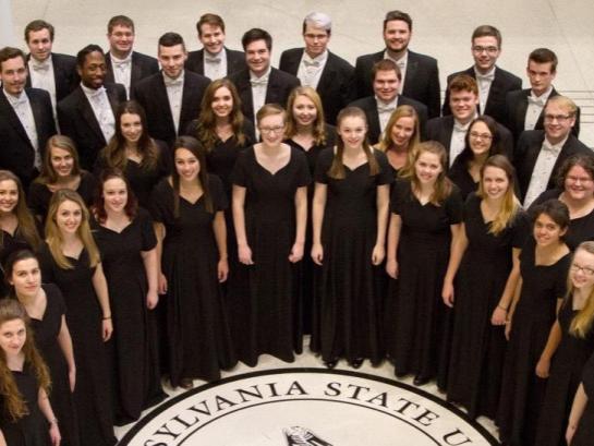 Members of the Penn State Concert Choir, wearing black tuxes and dresses, stand in a semi-circle and look up to smile for a photographer.