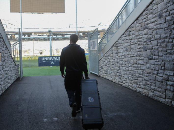 Male student rolls equipment case behind him as he looks on to an empty football stadium.