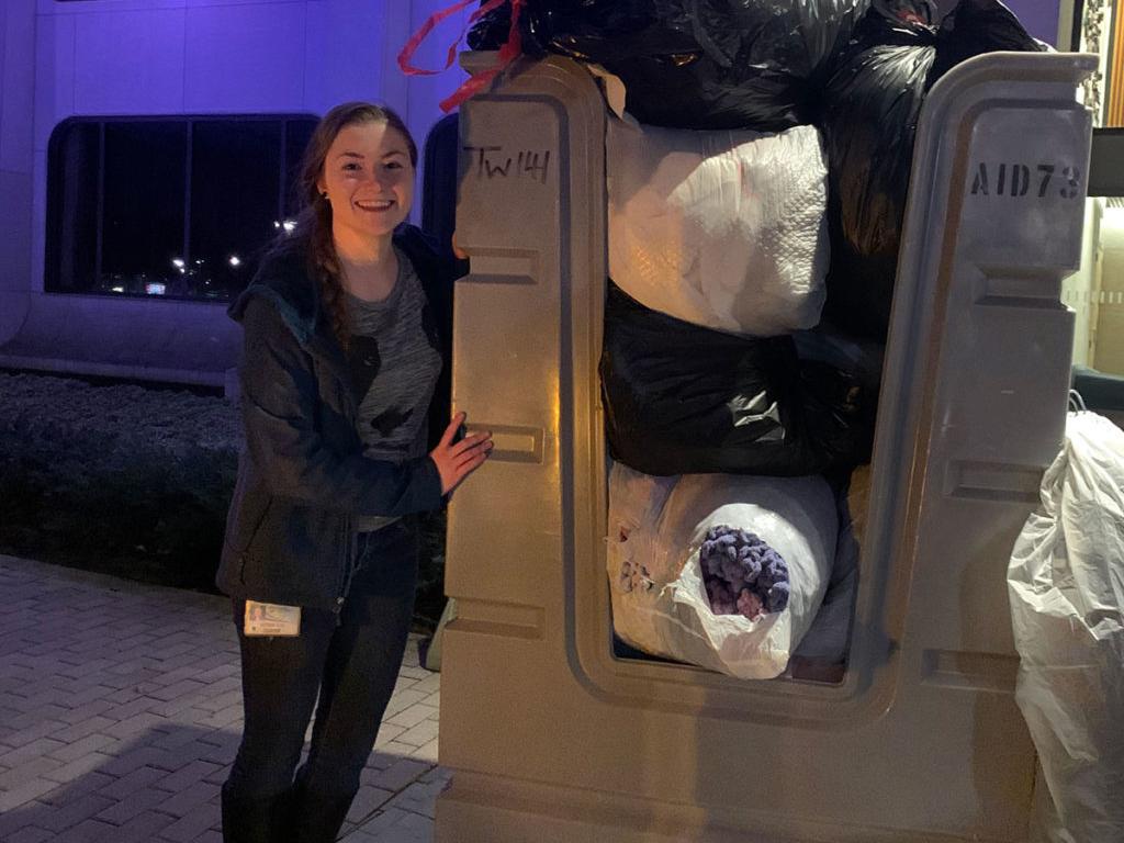 A young woman stands next to a large bin full of bags of winter coats.