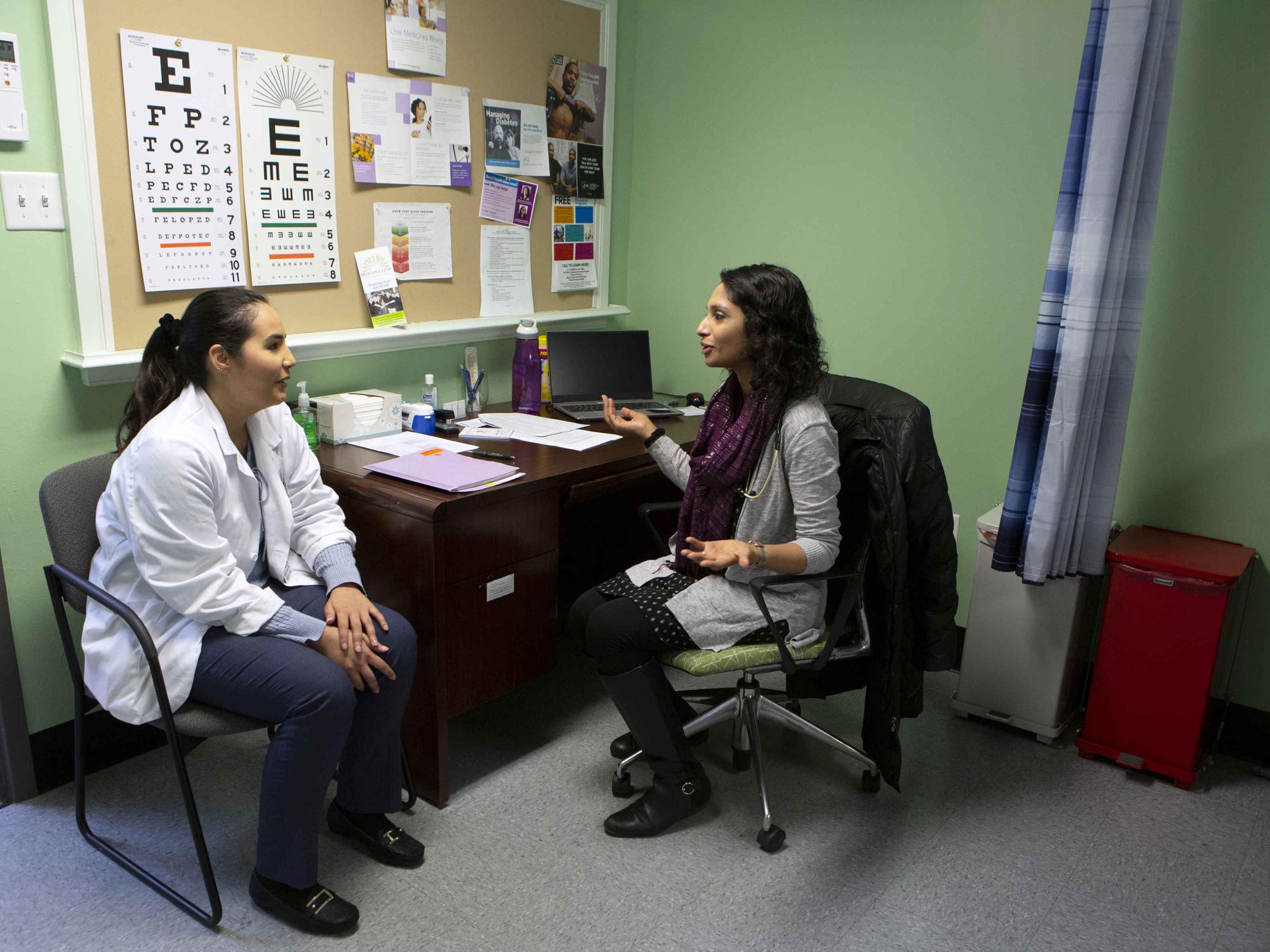 Dr. Leesha Helm, seated at a desk, gestures toward Penn State College of Medicine third-year student Maria Escudero during a shadowing visit at Beacon Clinic. A bulletin board with an eye chart and other papers on it is on the wall behind the desk.