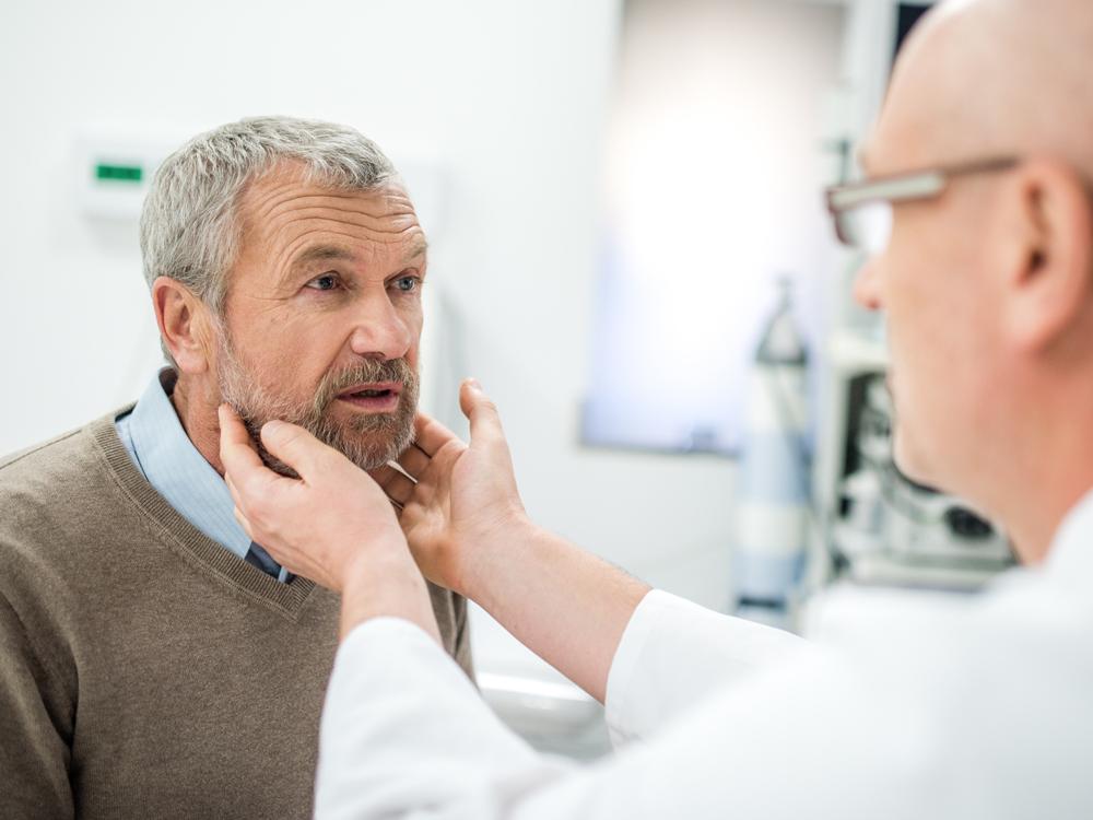 A male physician extends his hands toward the face of a male patient. Various medical equipment is in the background.