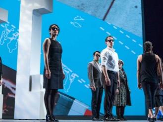 A small group of people stand rigidly in place while another group walks offstage.