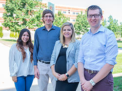 Four people stand on a sidewalk on a sunny day and smile. 