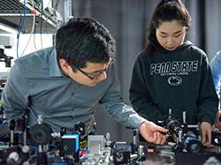 A young woman in a Penn State sweatshirt and a man in glasses and a button-down shirt stand near a large table with equipment and adjust the equipment 