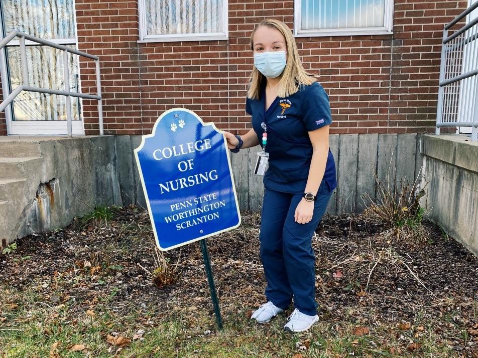 Logan DeSanto posing in nursing scrubs with College of Nursing sign