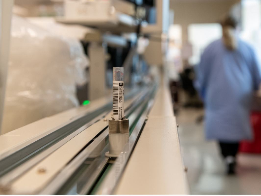 A vial is propped on a conveyor belt. In the background, someone in a lab coat is walking away out of focus.