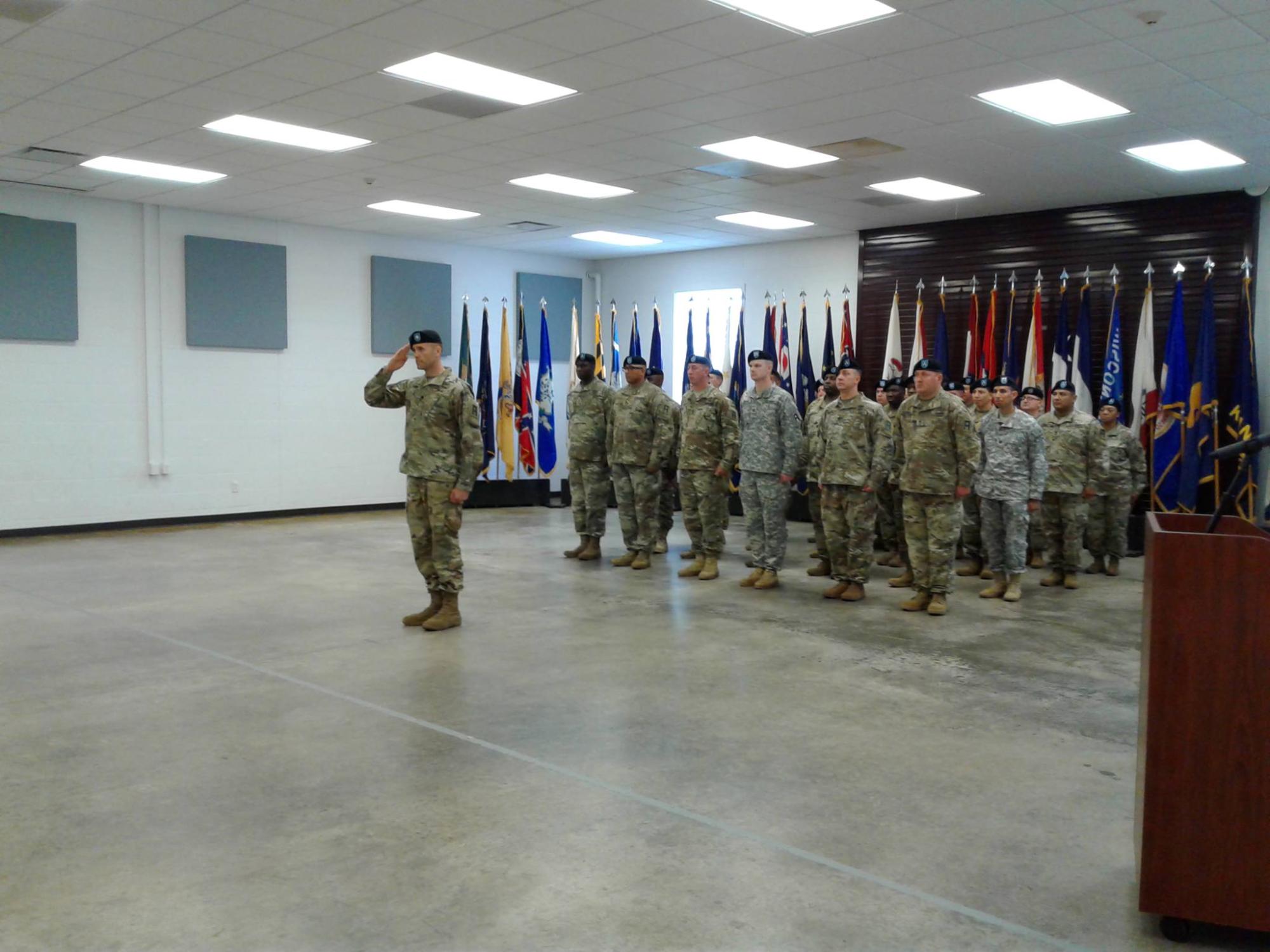 Saluting military man, dressed in uniform, stands in large room with group of soldiers in line behind him.