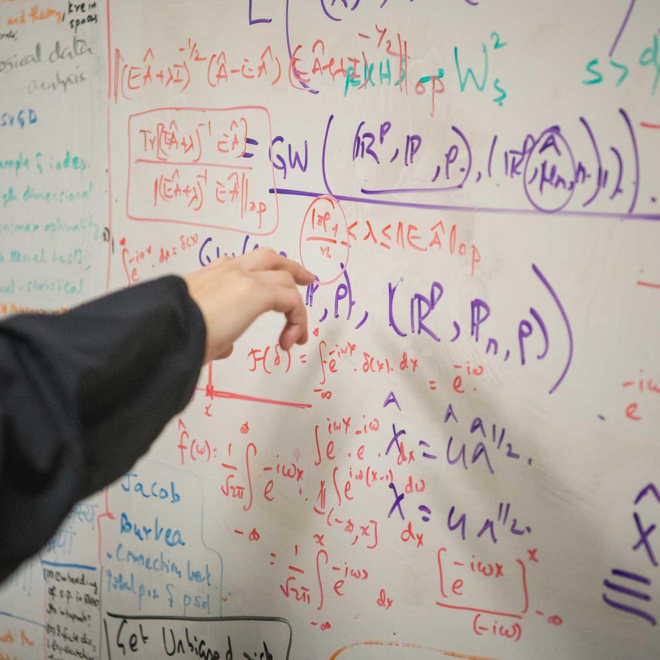 Hand points to white board that is full of equations written in different colors