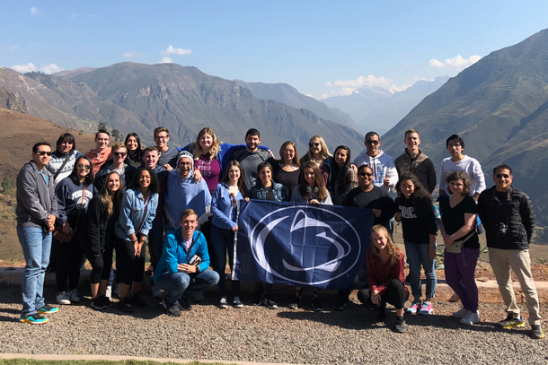 A group of 27 students and one professor pose in front of a mountain range while holding a Penn State flag. 