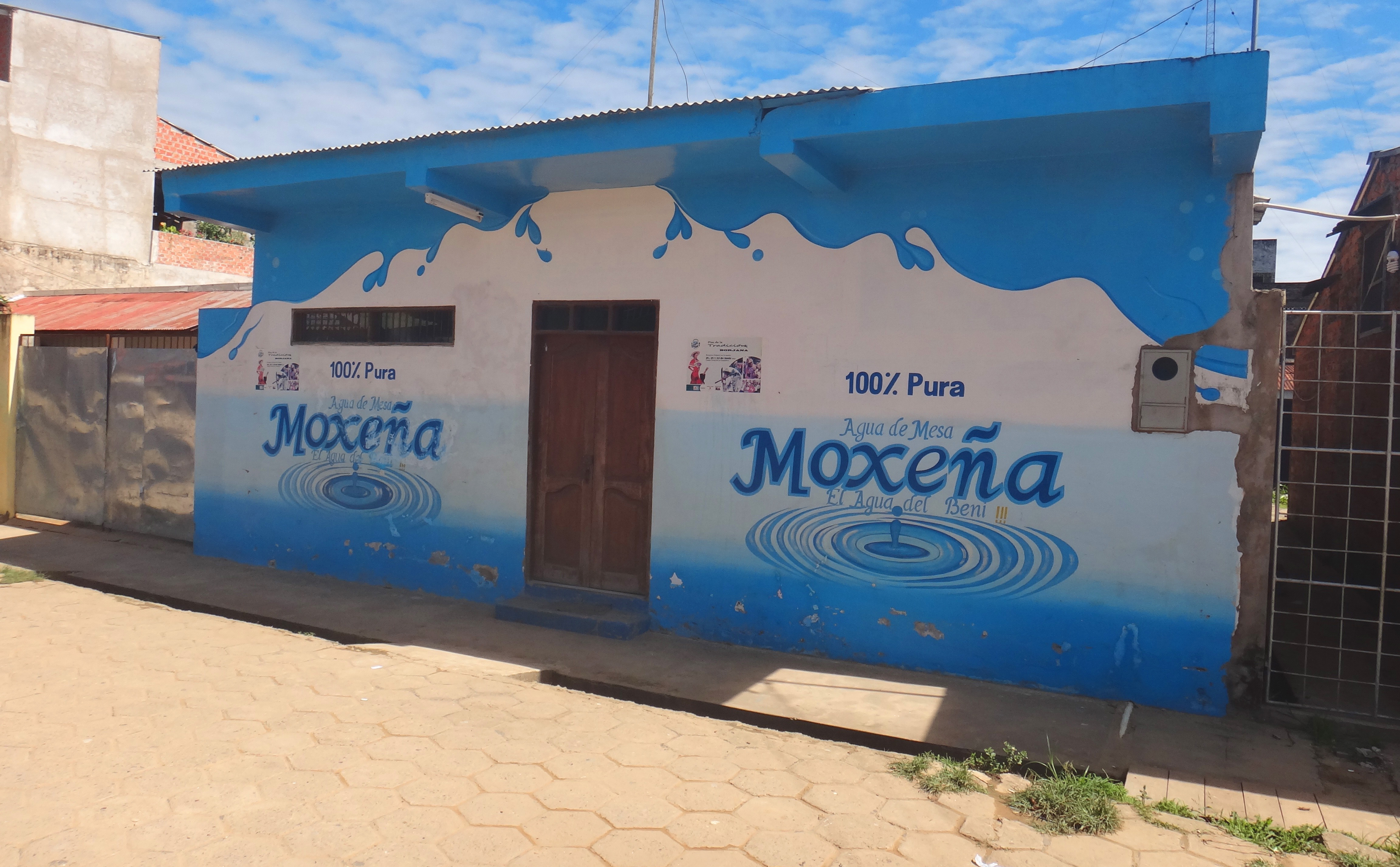 A small shop that sells water on a dirt road situated among other buildings along a dusty, unpaved road