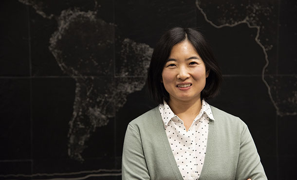 A woman in a white button down with polka dots and a pale green sweater poses in front of a world map.